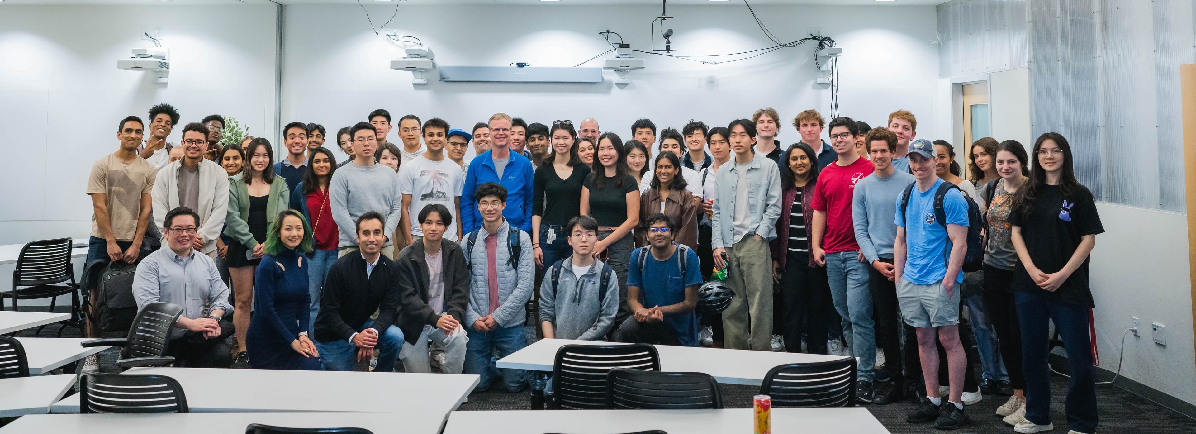 A large group of young adults, appearing to be students, are posing for a photo in a classroom or lecture hall. They are standing and sitting in rows, with many smiling and looking at the camera. The room has whiteboards and projectors visible.