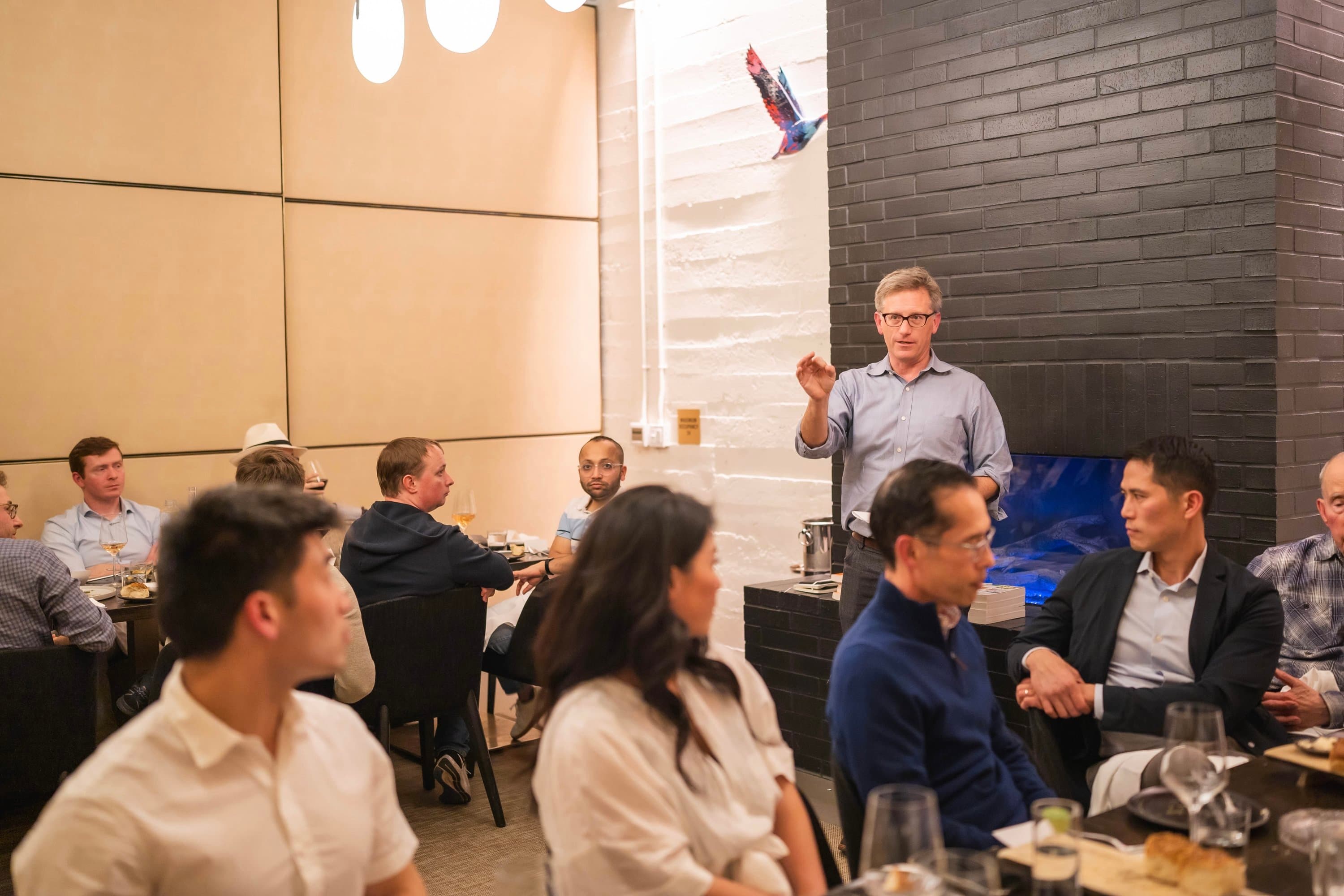 A man in a blue shirt and glasses stands and gestures while speaking to a group of people seated at tables in a restaurant.