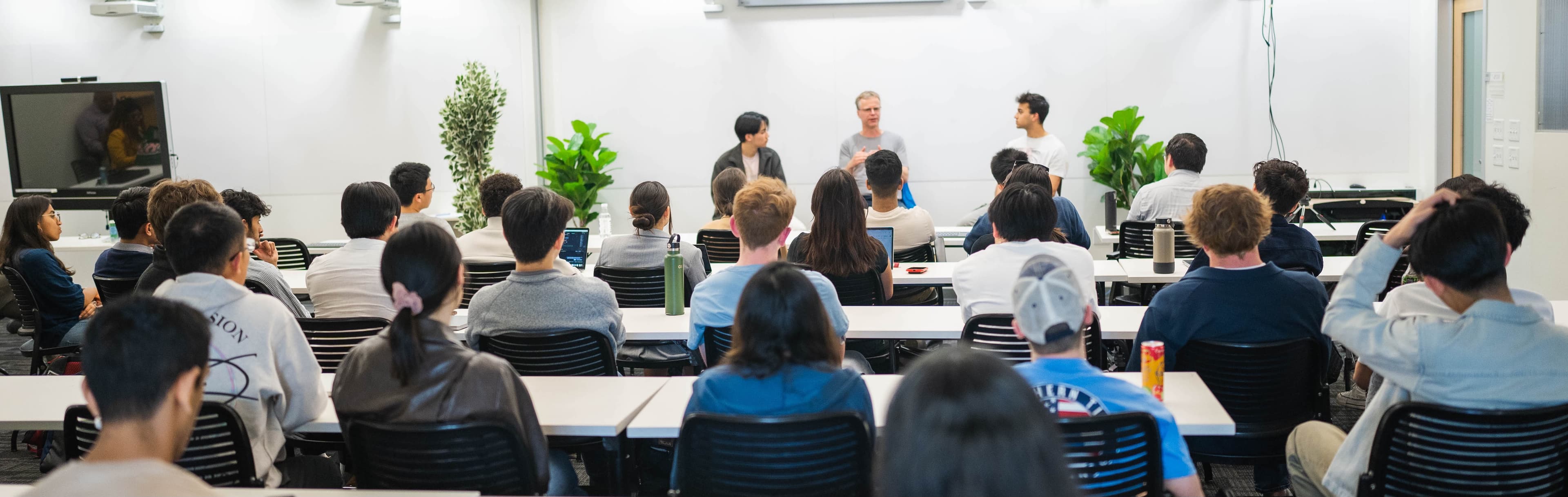 A group of people are seated in a classroom or lecture hall, facing forward. Two individuals are standing at the front, appearing to present or lead a discussion. Plants are visible in the background, and a large screen is mounted on the wall.