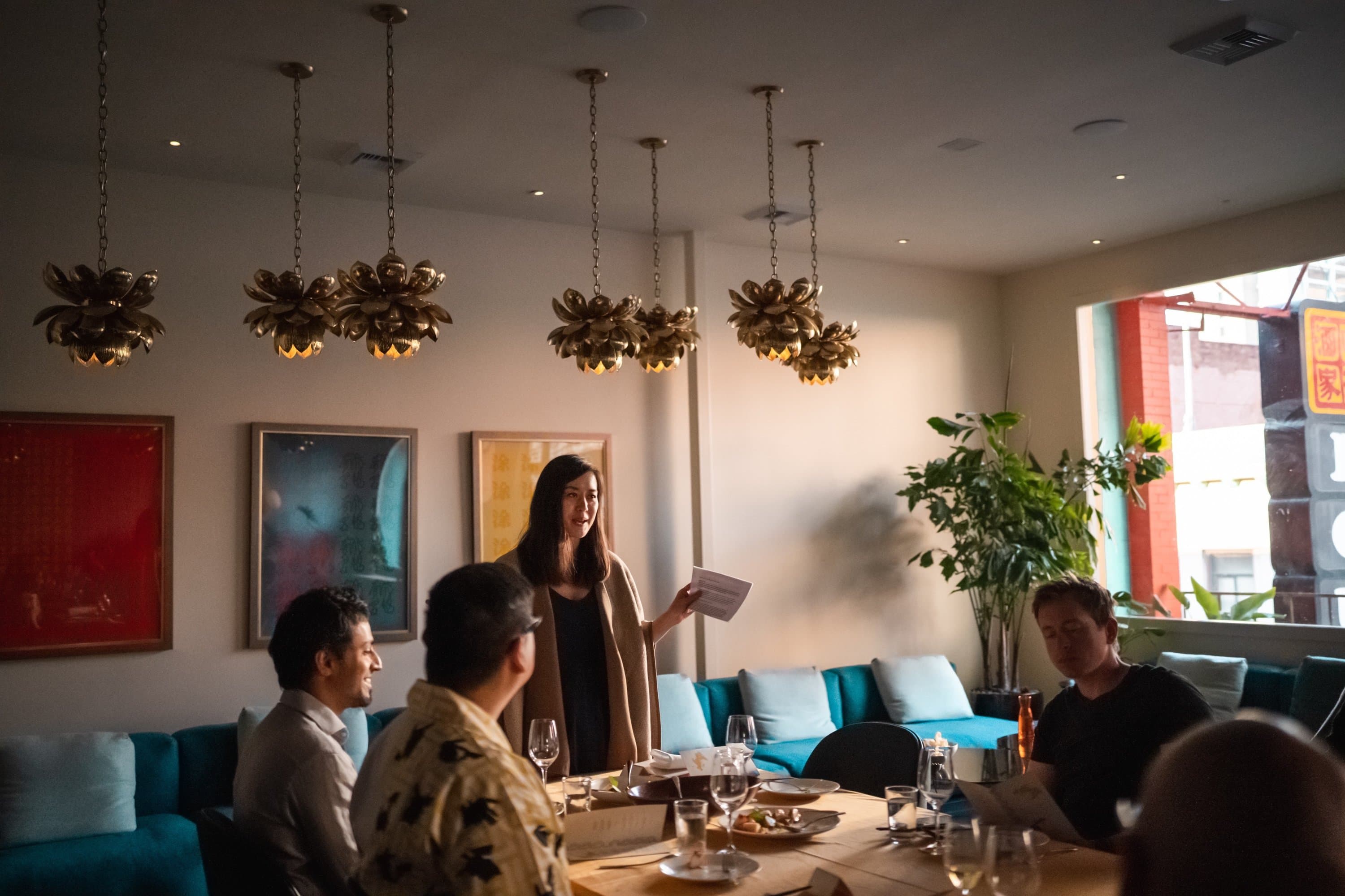 A woman stands at the head of a table in a restaurant, holding papers and speaking to a group of people seated around the table. The room is decorated with modern art and has several ornate pendant lights hanging from the ceiling.