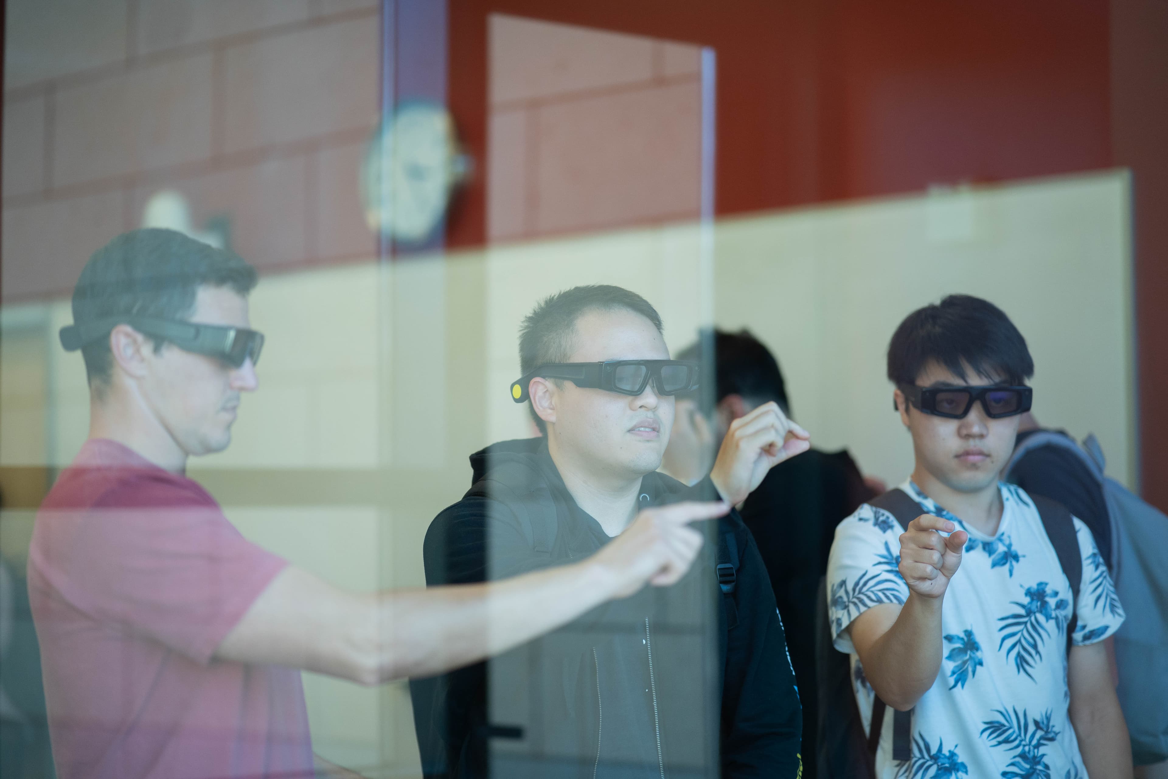 Three men wearing black 3D glasses are looking forward and pointing. The man on the left wears a pink shirt, the middle man wears a black hoodie, and the man on the right wears a white t-shirt with a blue floral pattern.