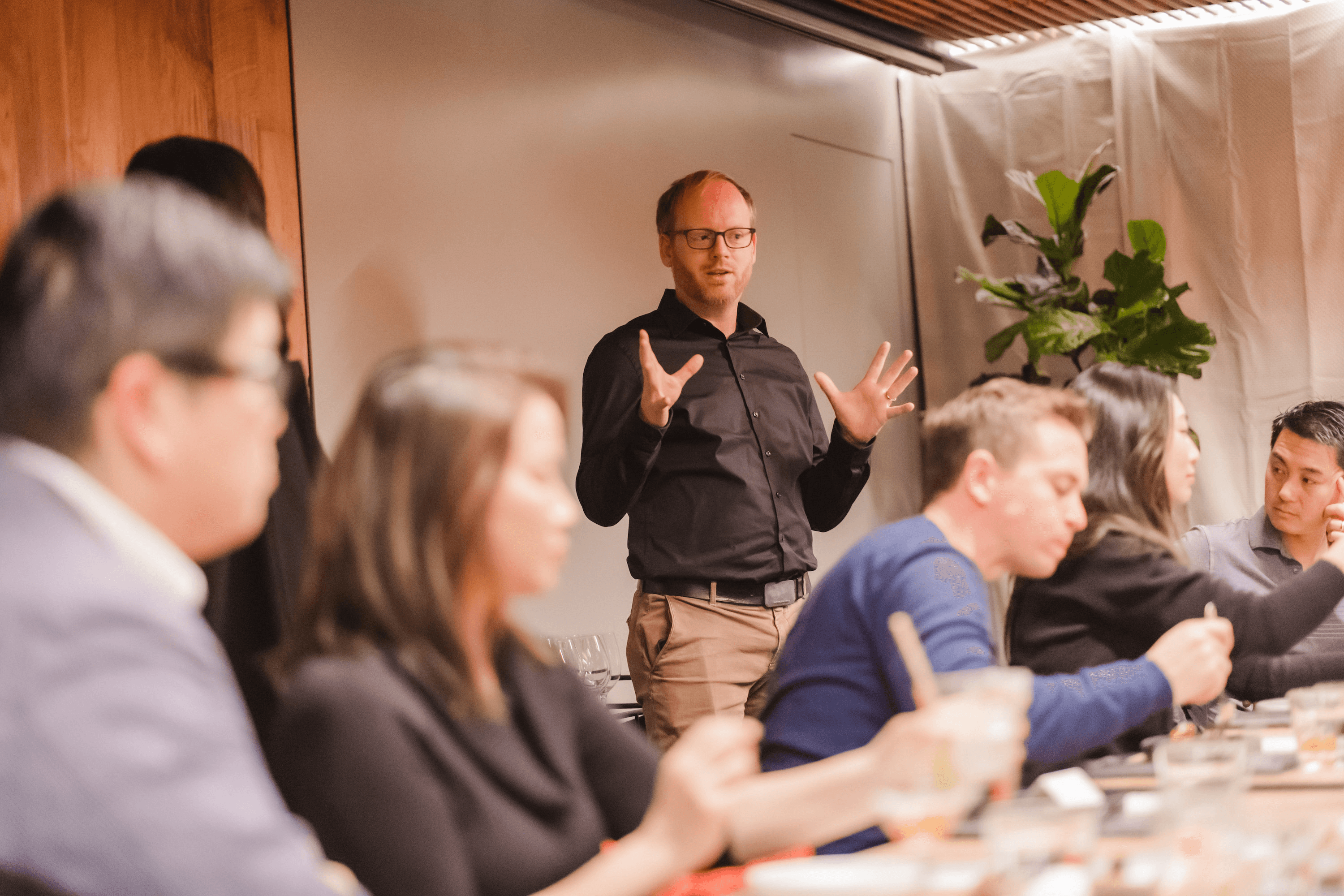 A man with glasses and a black shirt stands and gestures with his hands while speaking to a group of people seated at a table.